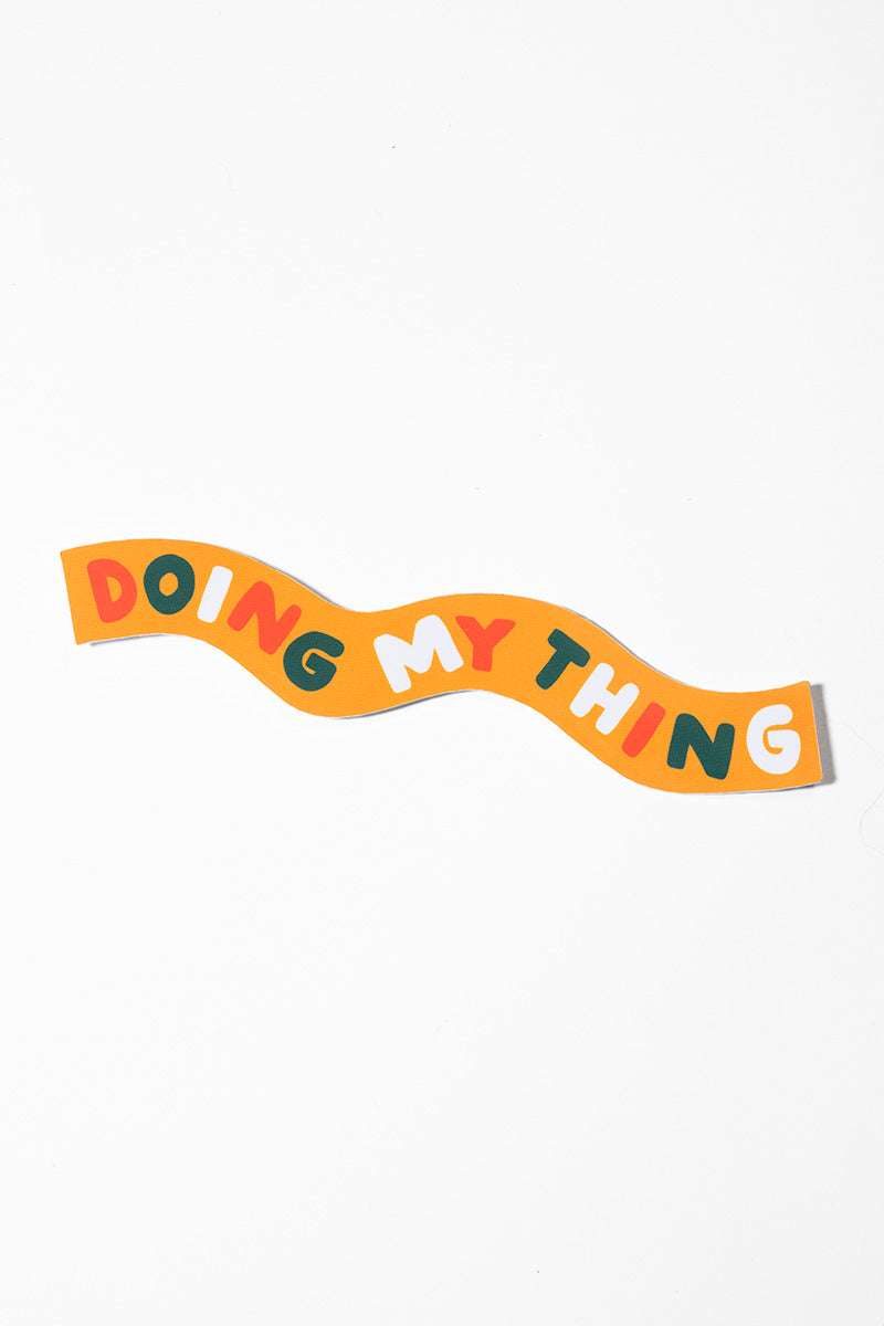 Sticker that reads "doing my thing" in wavy green, orange, and white lettering on a wavy yellow background all shot on a white background.