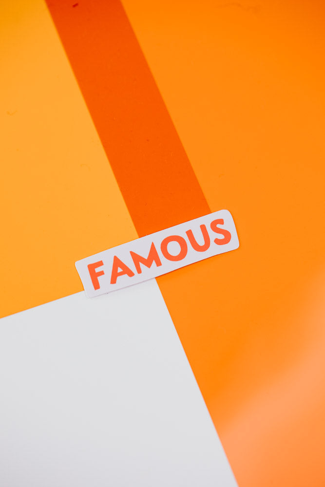 An image showing a single white rectangular sticker with the word "FAMOUS" written in large red letters, placed against a vibrant orange background.