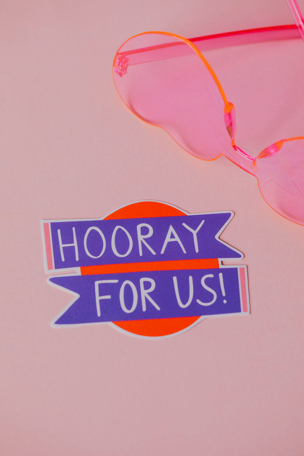 The image shows a close-up of a pink and orange glass object, likely a drinking glass or wine glass. In the background, there is a sticker that says "HOORAY FOR US!" in bold, purple text against an orange background.