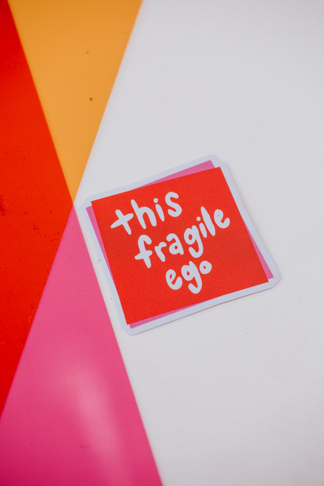  single square sticker with white text reading 'this fragile ego' on a bright red background with pink borders, photographed against a geometric background of white, orange, and pink sections