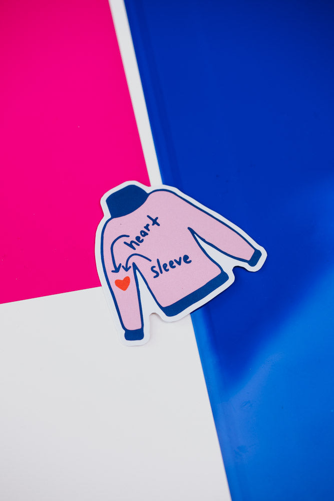 Pink sweater sticker labeled "heart" and "sleeve" with an arrow pointing to a red heart on the left sleeve, placed on a bright pink and blue background.