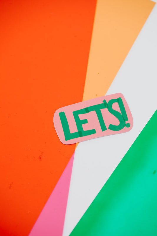 The image shows a close-up of a pink and green "Let's!" sticker on a vibrant orange and yellow background.