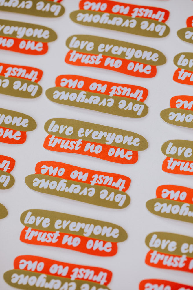 Multiple stickers with the text "love everyone trust no one" in gold and orange, arranged on a white surface.