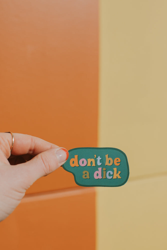 A hand holding a sage green speech bubble sticker with multicolored lettering against an orange and beige gradient background
