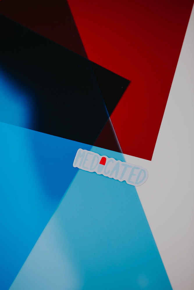 An abstract image featuring geometric shapes in various shades of blue, black, and red, with a white "MEDICATED" sticker or label prominently displayed in the foreground.