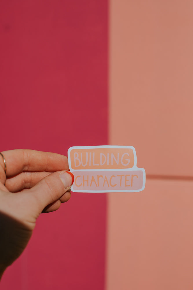 A hand holding a peach-colored sticker with 'Building Character' text against a pink and coral gradient background