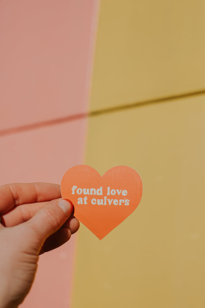 A hand holding an orange heart-shaped sticker with retro-style text reading 'found love at culvers' against a split background of peachy pink and warm yellow