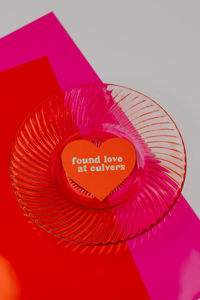 An orange heart sticker reading 'found love at culvers' placed on a clear glass plate with radial ridges, set against a backdrop of red and hot pink geometric shapes