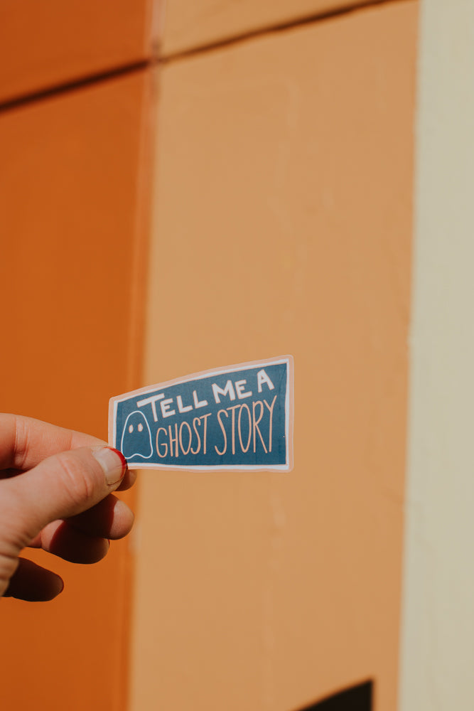 
A close-up image of a hand holding a small rectangular sticker with the text "Tell me a ghost story" in playful, handwritten style. The sticker has a simple ghost icon next to the text on a blue background. The background behind the hand is a warm, two-tone wall in shades of orange and cream.